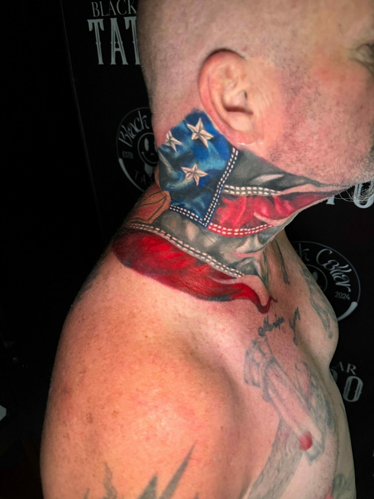 Close-up of a person's neck and shoulder with tattoos, showing a red mark or injury, and a flag tattoo on the neck.