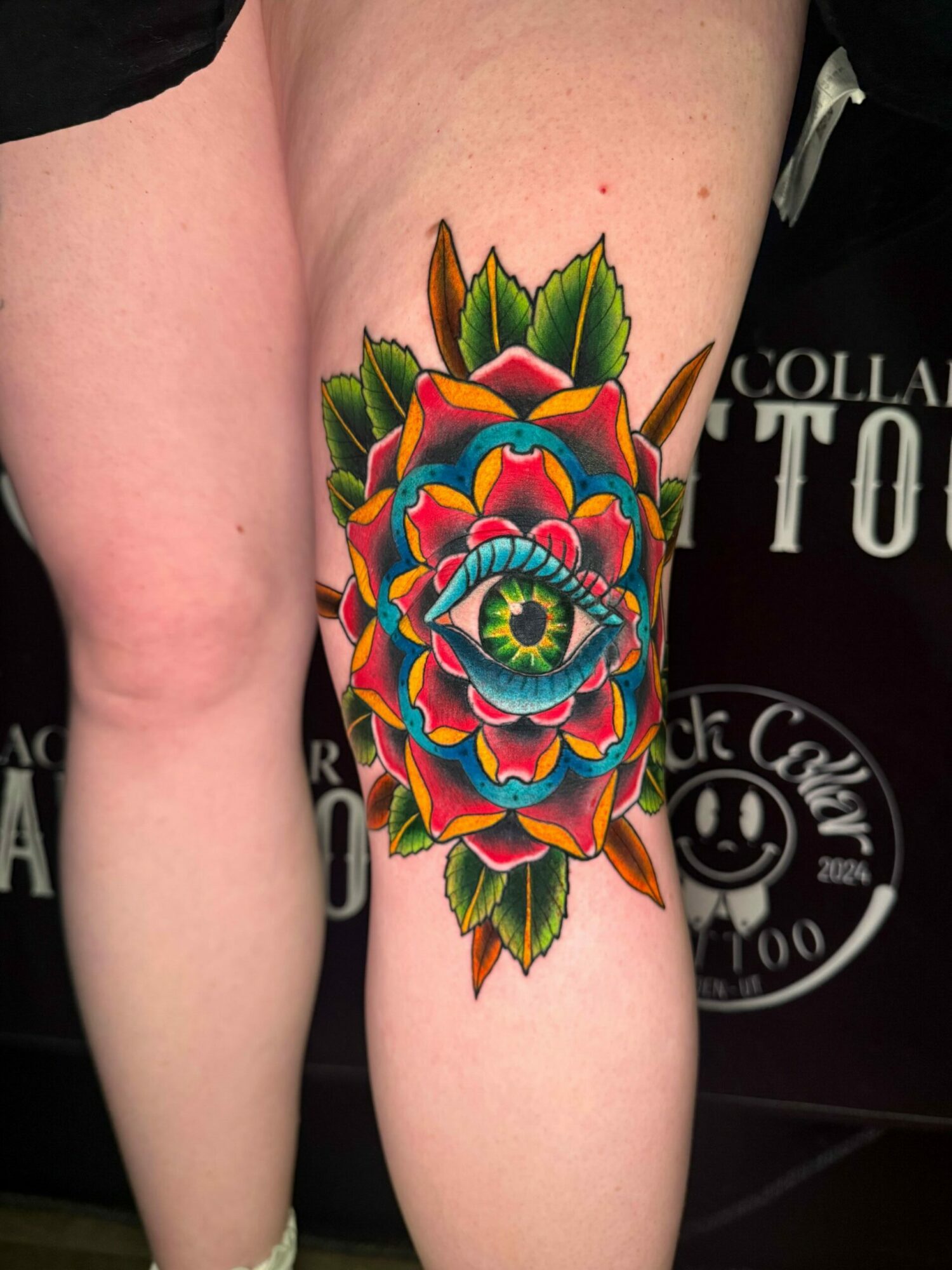 Colorful floral tattoo on arm with green leaves, vibrant petals, and a central eye, in traditional style.