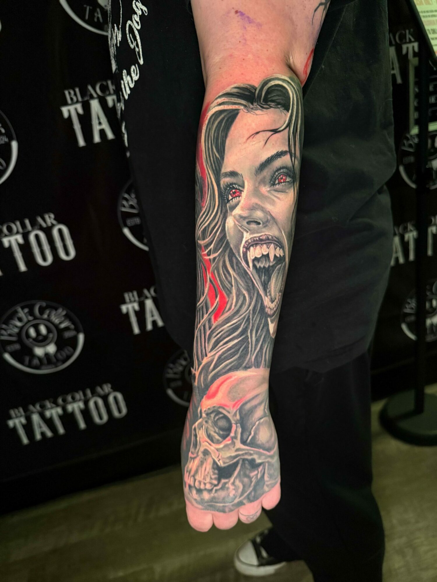 Tattooed arm with a detailed face, skull, and abstract designs, extending from wrist to elbow, in black and red ink.