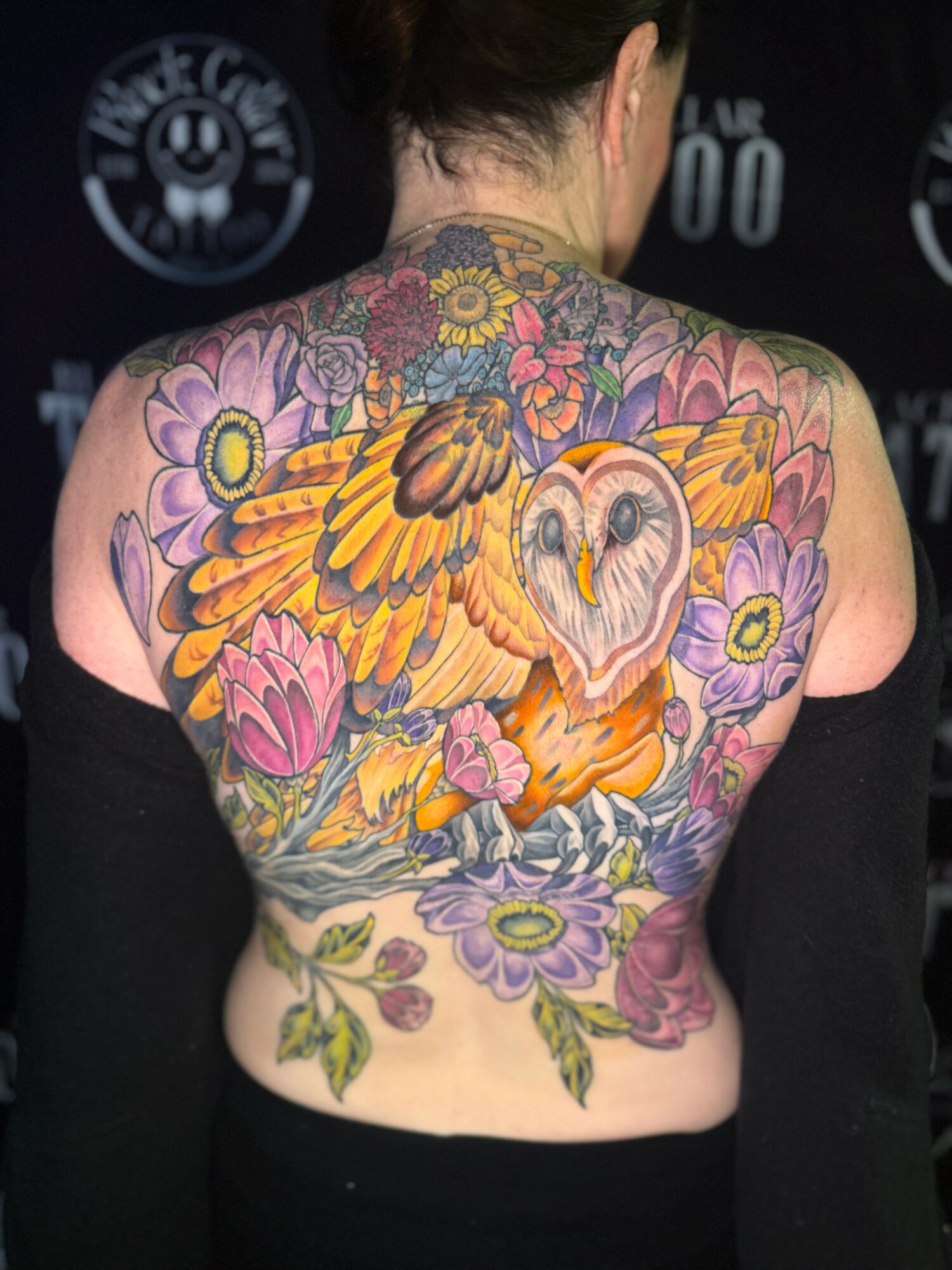 Back view of a person with a colorful tattoo of a swan, flowers, and leaves covering the back and shoulders.