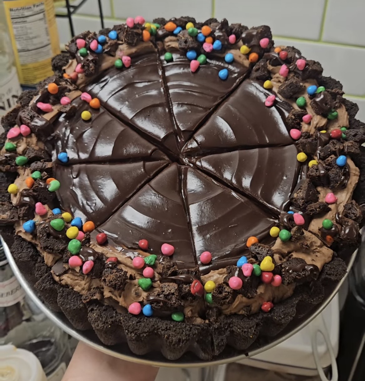Chocolate cake with colorful sprinkles, sliced into pieces, on a white plate, with a kitchen background.