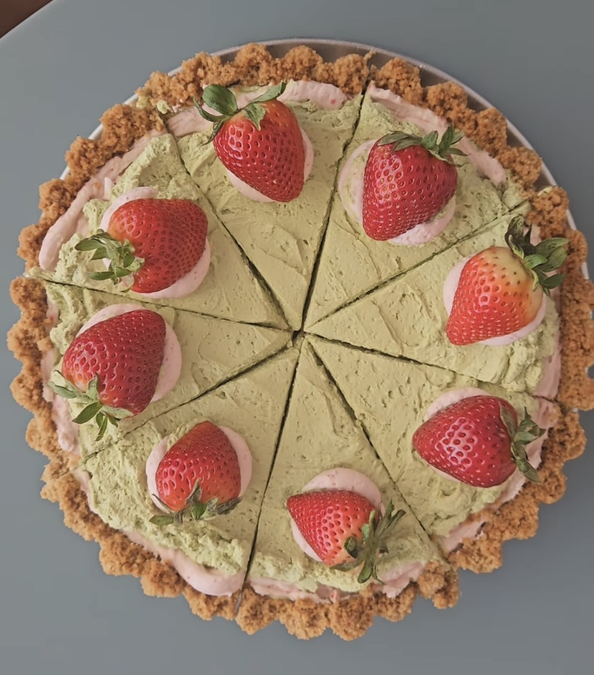 A round cheesecake topped with strawberries and mint leaves, sliced into eight pieces, with a crumbly crust, on a blue surface.