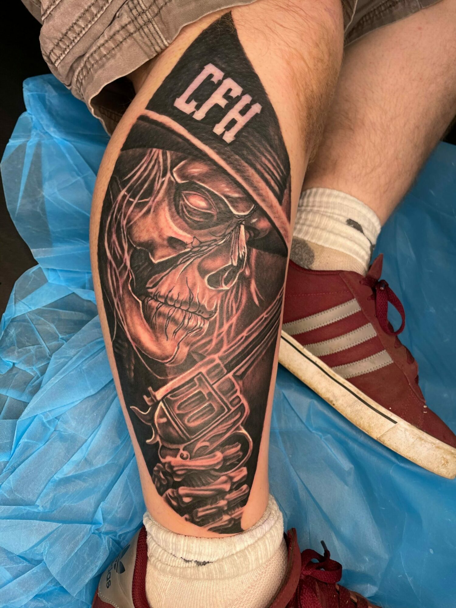 Close-up of a person's arm with a detailed black and gray tattoo of a skull wearing glasses, with the word 'TECH' above, on a blue surface.