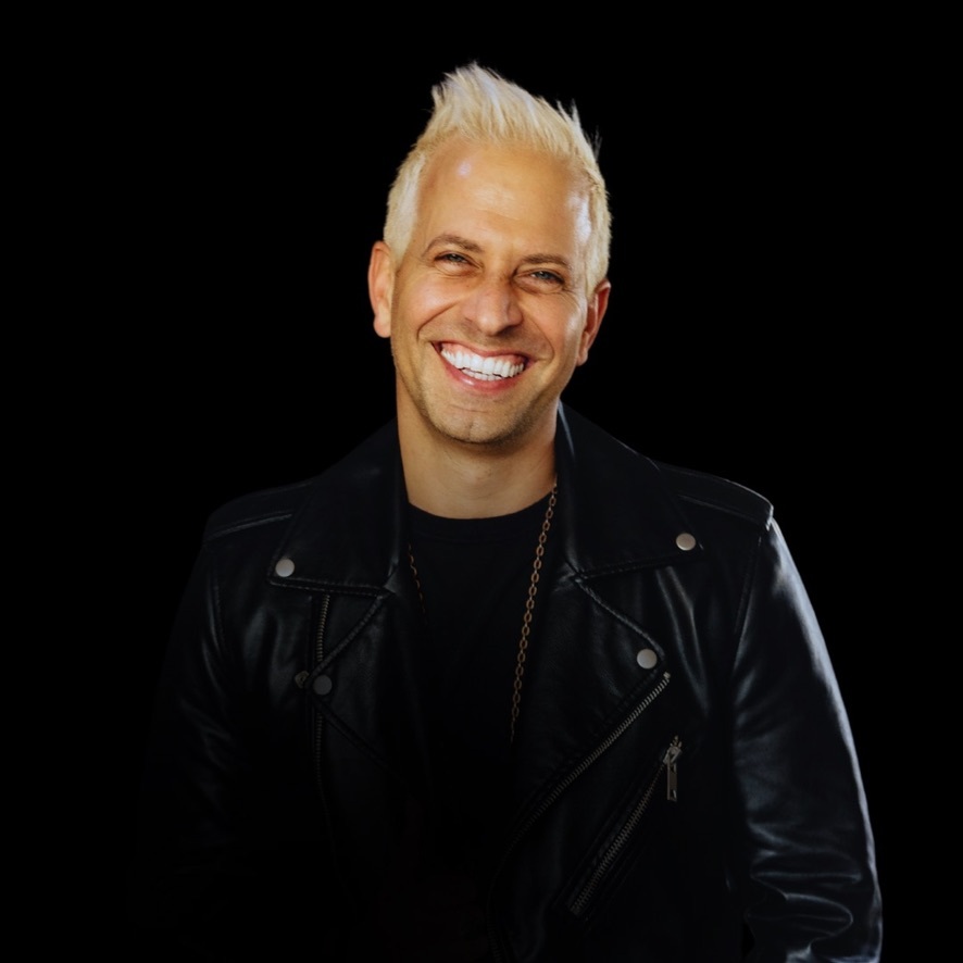 Smiling person with short, platinum blonde hair wearing a black leather jacket against a black background.