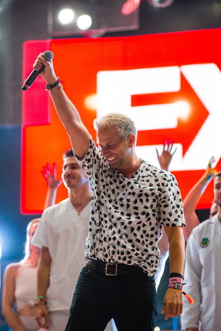 Man with short blond hair, wearing a black and white patterned shirt, holding a microphone and smiling, with people in the background.