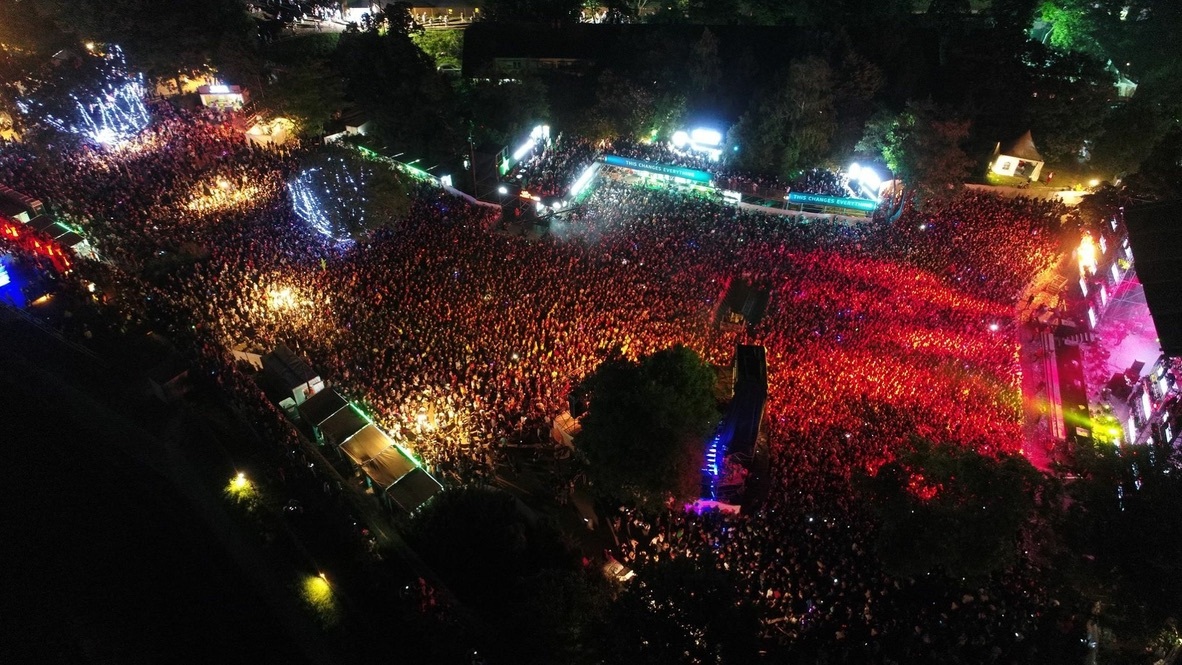 Large crowd at night outdoor event with colorful stage lights and illuminated area, surrounded by trees and dark sky.