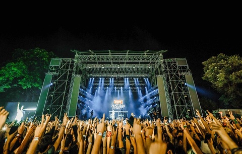Concert stage with bright lights and a crowd raising hands at night, trees visible on sides.