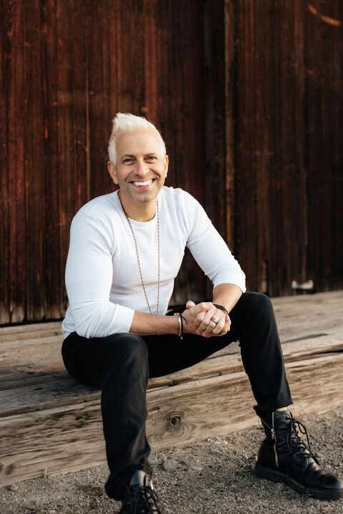 Person with short white hair smiling, sitting outdoors against a wooden wall, wearing a white long-sleeve shirt and black pants.