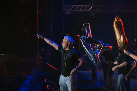 Person with blue hair pointing to the left on stage with musical instruments and performers in background