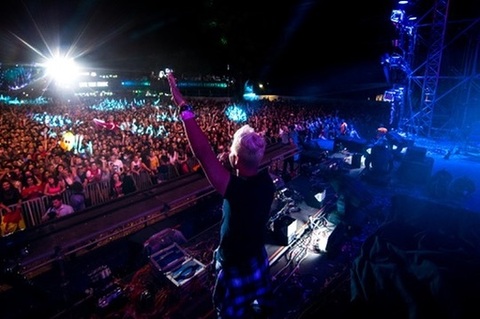 DJ with raised arms performs on stage at a large concert with a crowd and bright lights.