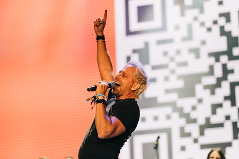 A man with gray hair singing into a microphone, raising one hand, on stage with a digital background.