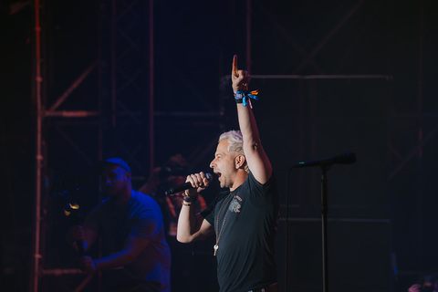 Person singing into microphone with one arm raised on stage, dark background, stage lighting, and other people in the background.