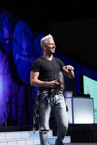 Man with white hair smiling on stage, wearing a black t-shirt and jeans, holding a device, with a blue-lit background.