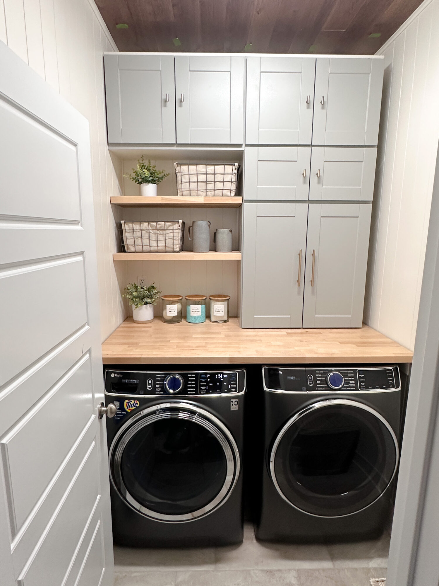 Laundry room with washing machine and dryer, shelves with baskets and plants, and cabinets above.