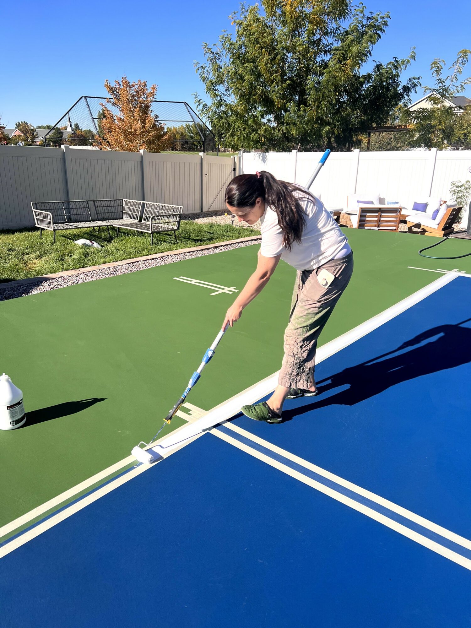Person cleaning a tennis court with a long-handled tool, outdoors on a sunny day, with trees and a fence in background.