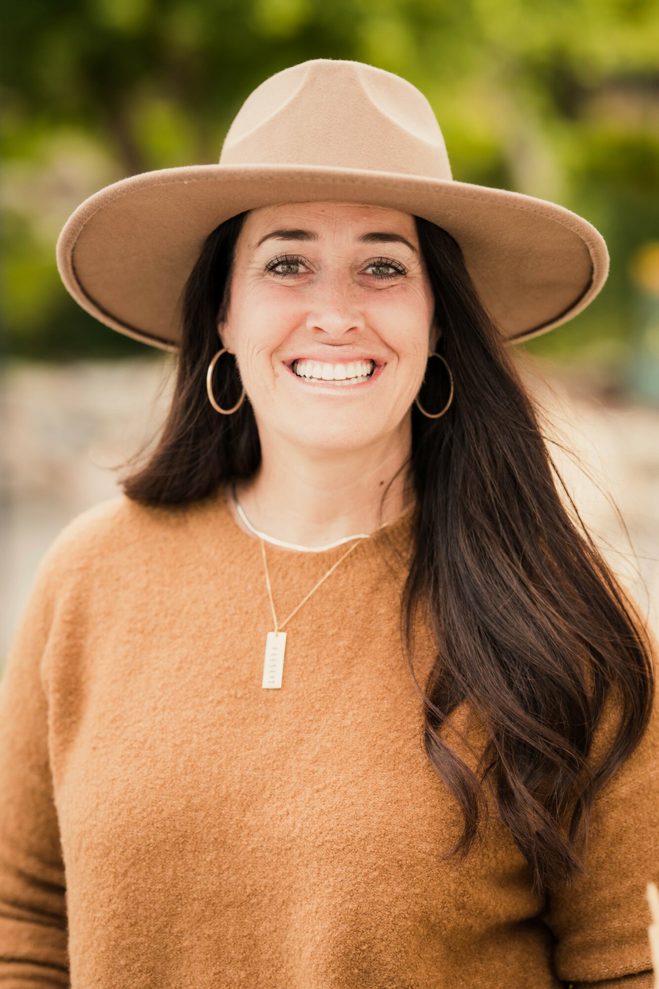 Smiling woman wearing a wide-brimmed hat, hoop earrings, and a brown sweater outdoors with greenery in background.