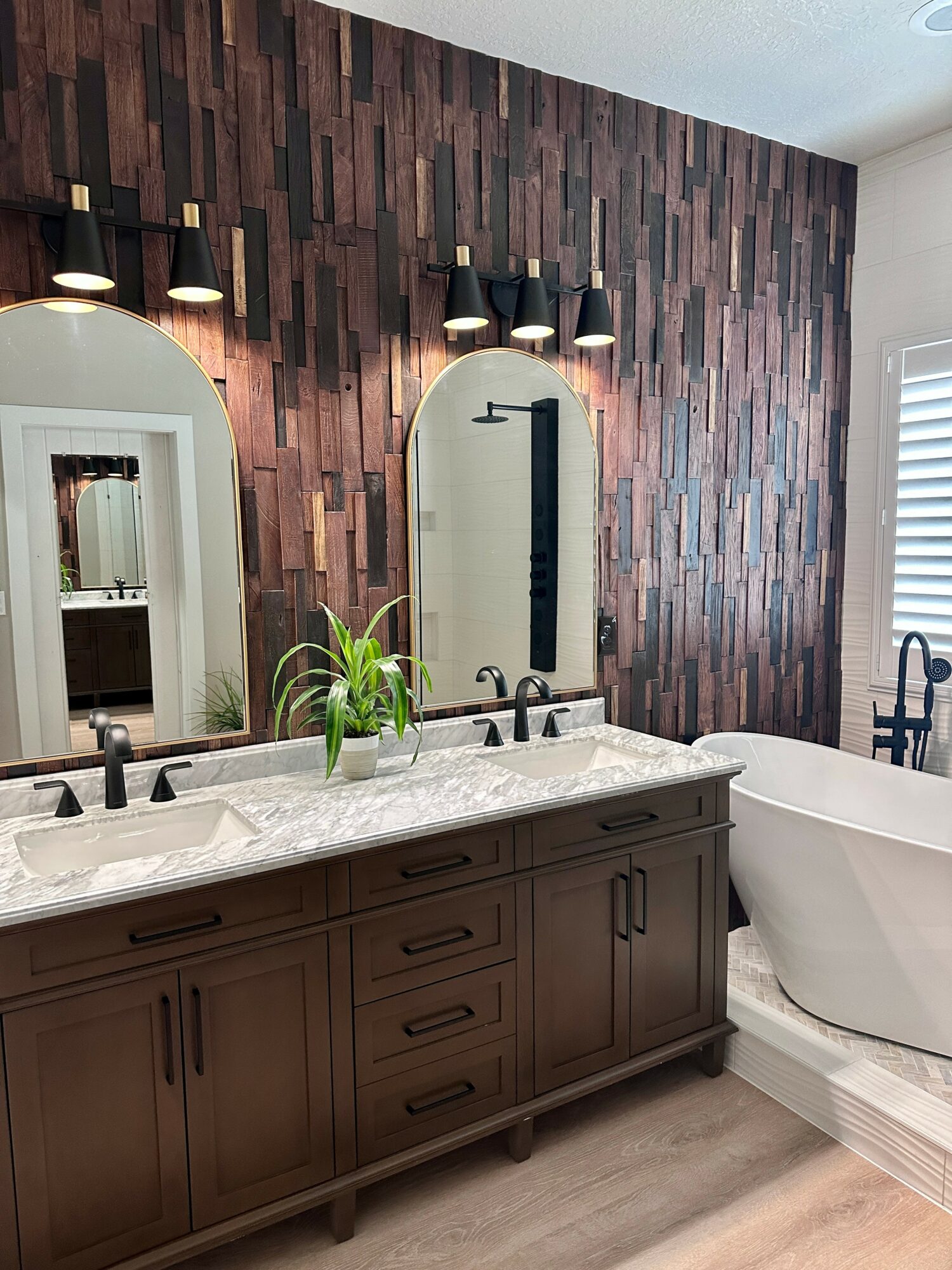 Bathroom vanity with two mirrors, a plant, and a bathtub against a dark wood-paneled wall.