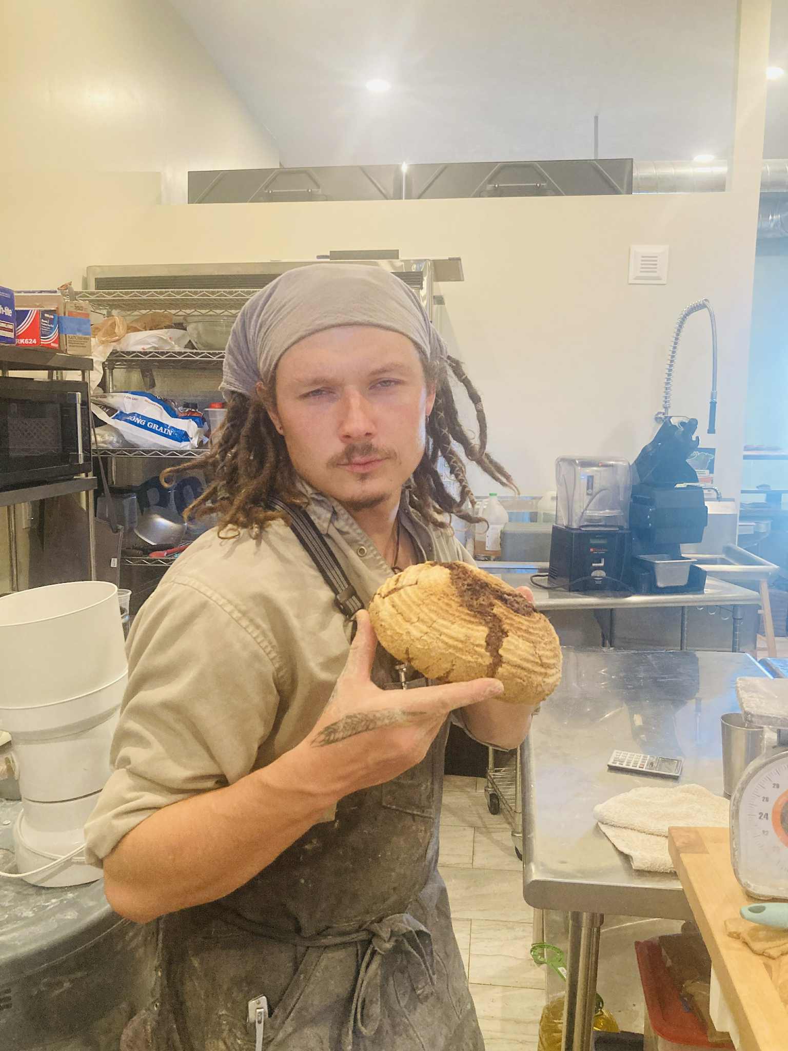 Person with dreadlocks and headscarf holding a large cookie in a kitchen setting.