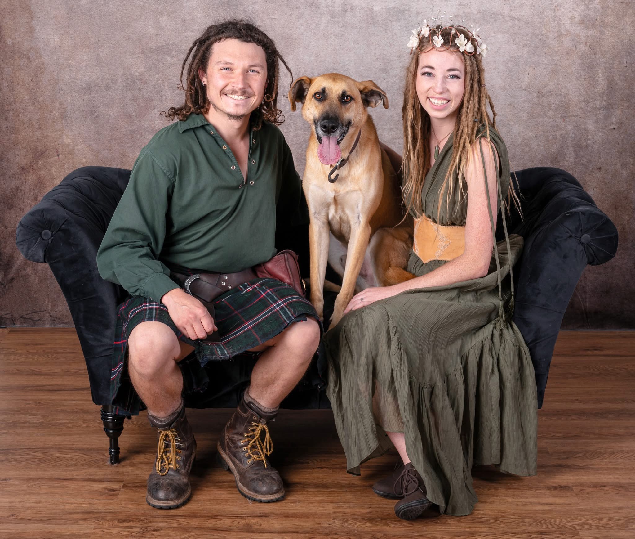 Two people and a dog sitting on a black sofa, smiling, with a plain background.