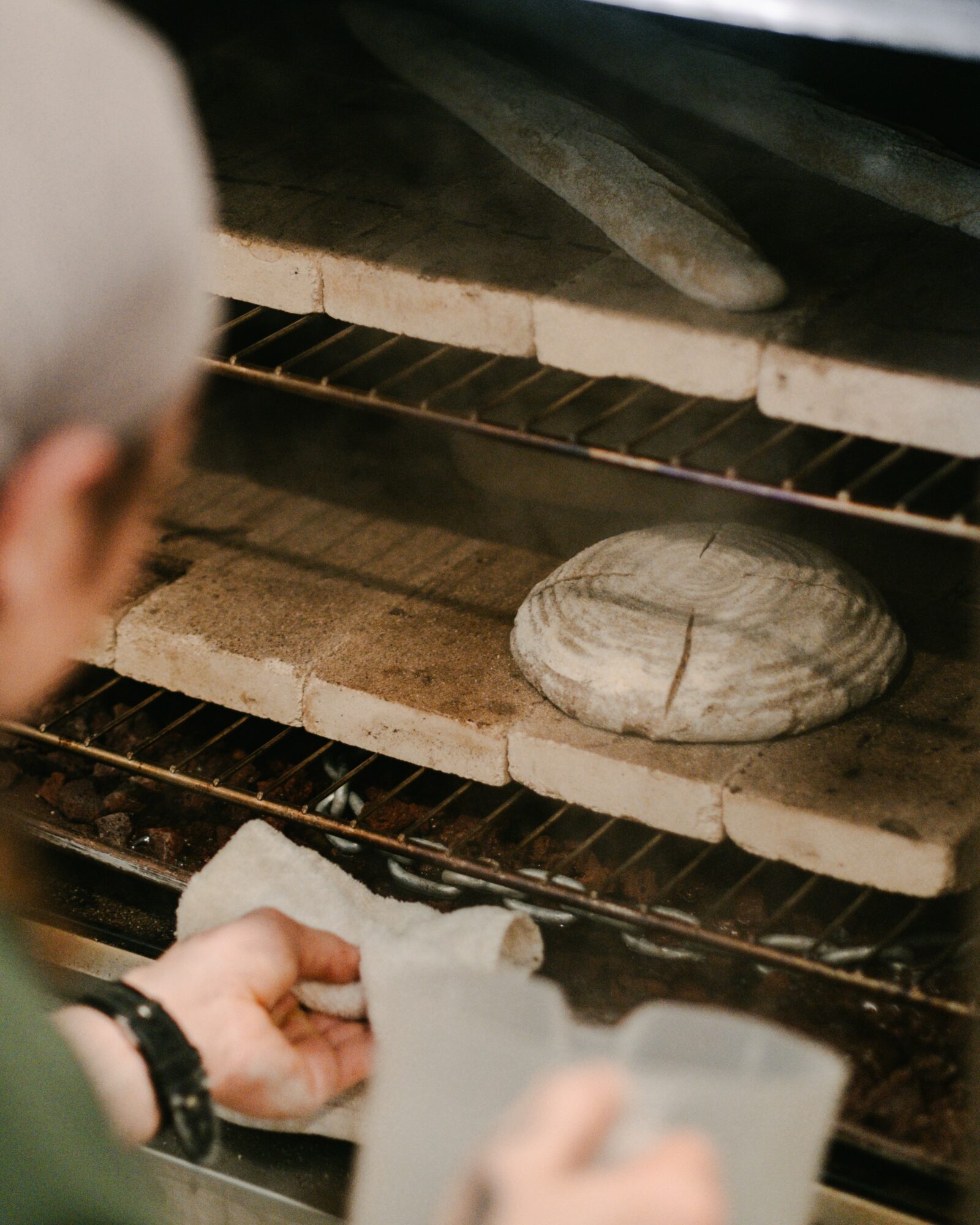 Person's hand holding paper towel near a round loaf of bread inside an oven with baking stones.