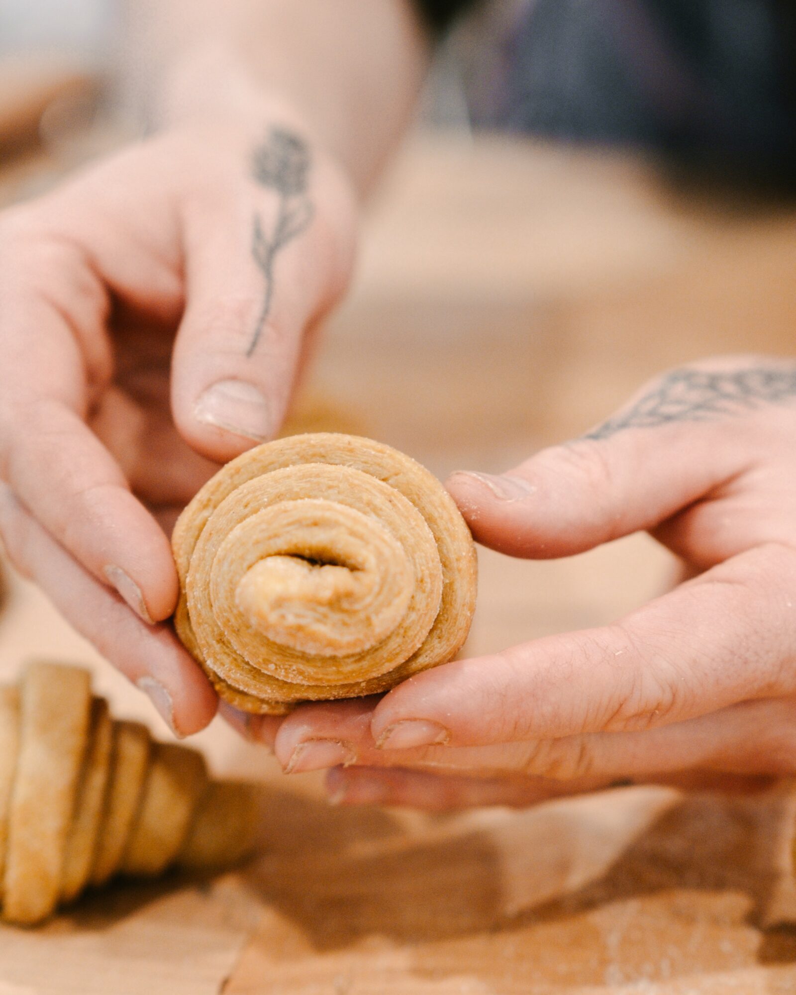 Two hands holding a cinnamon roll, with a tattooed finger visible, on a wooden surface.