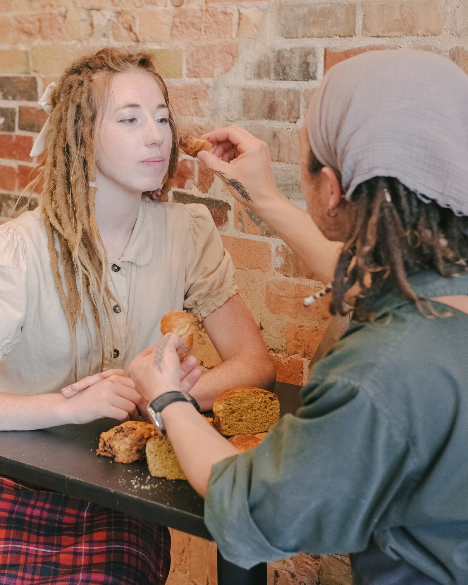 Person with gray headscarf applying makeup to young woman with dreadlocks, sitting at a table with food, brick wall background.