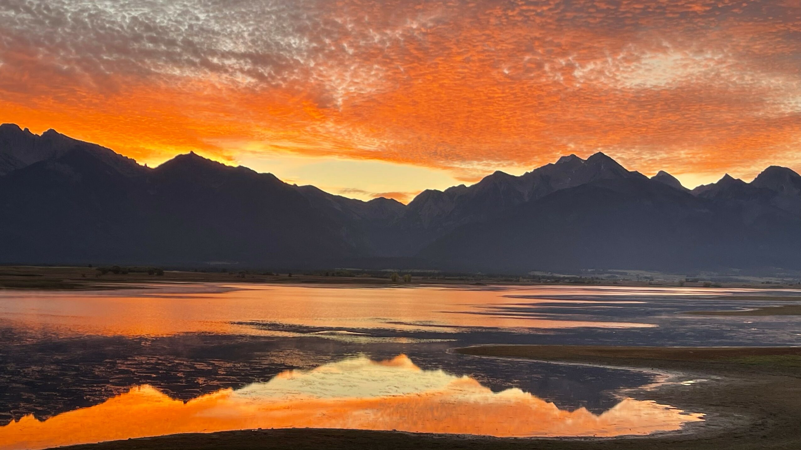 Sunset over mountains with reflected sky and water, vibrant orange and red hues, silhouetted mountain range, calm water surface.