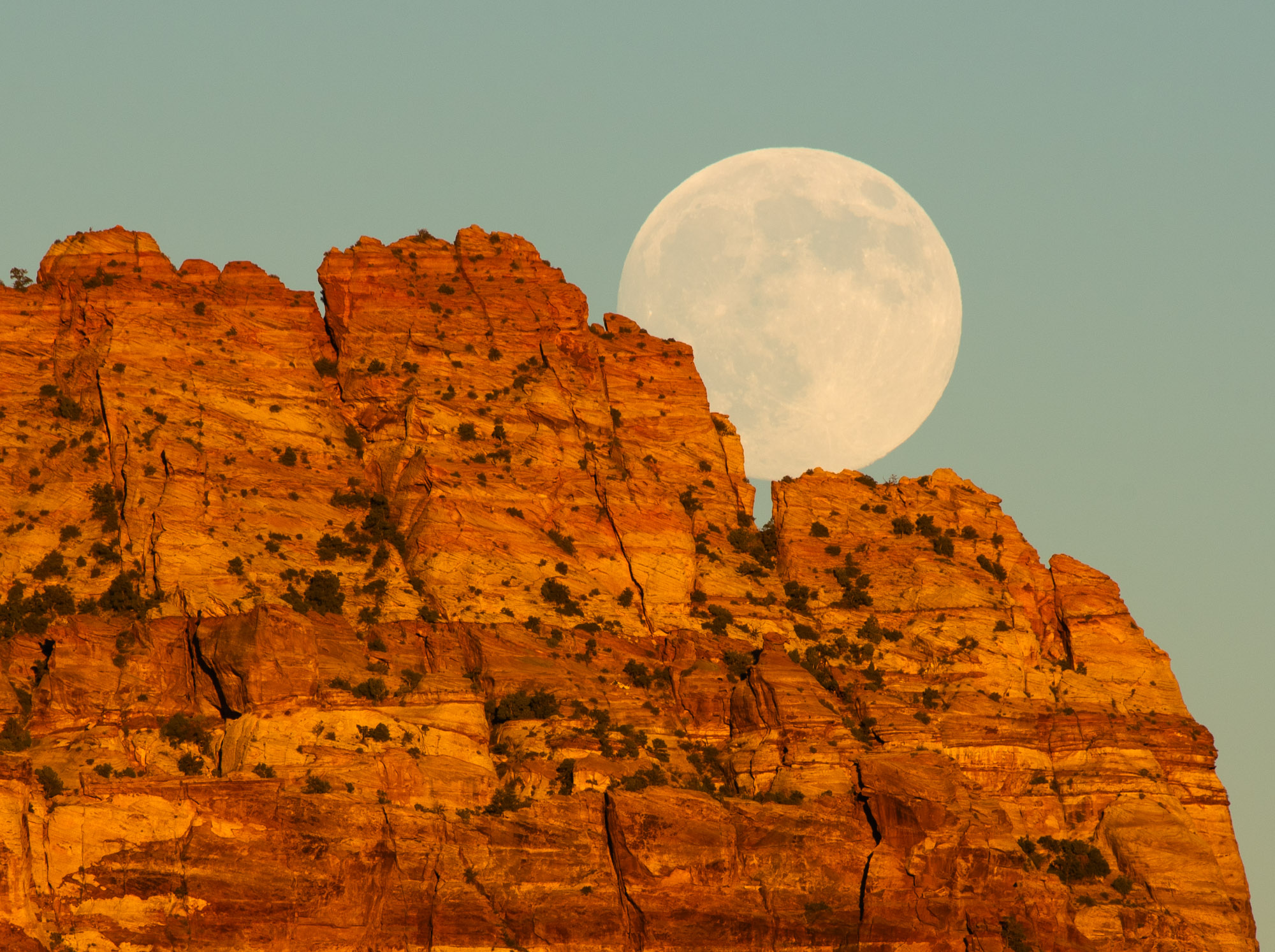 Full moon rising behind a rugged, reddish-brown mountain range with a clear sky.