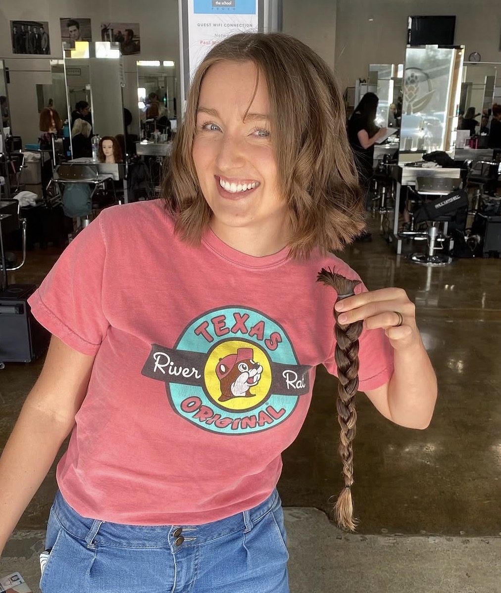 Woman with shoulder-length hair smiling, wearing a pink T-shirt with a graphic, holding a braid, in a salon. Word count: 20