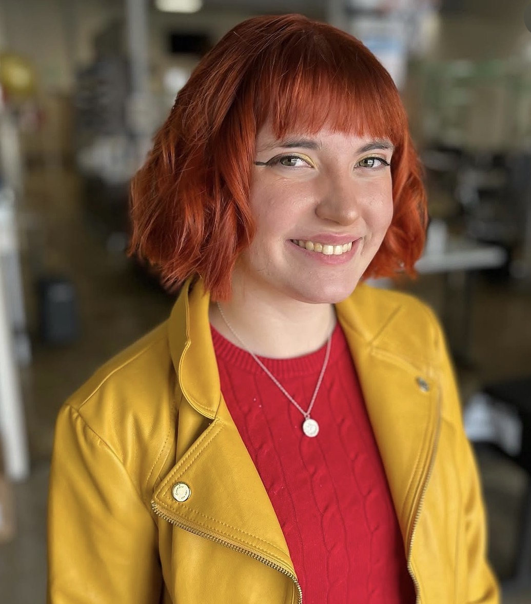 Smiling woman with red hair wearing a yellow jacket and red top, in an indoor setting.