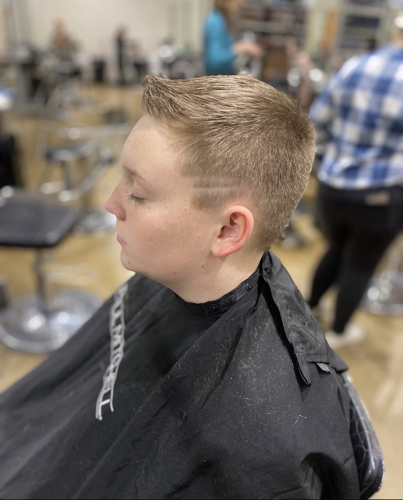 Young boy with short, styled hair getting a haircut in a salon, wearing a black cape, with blurred background.