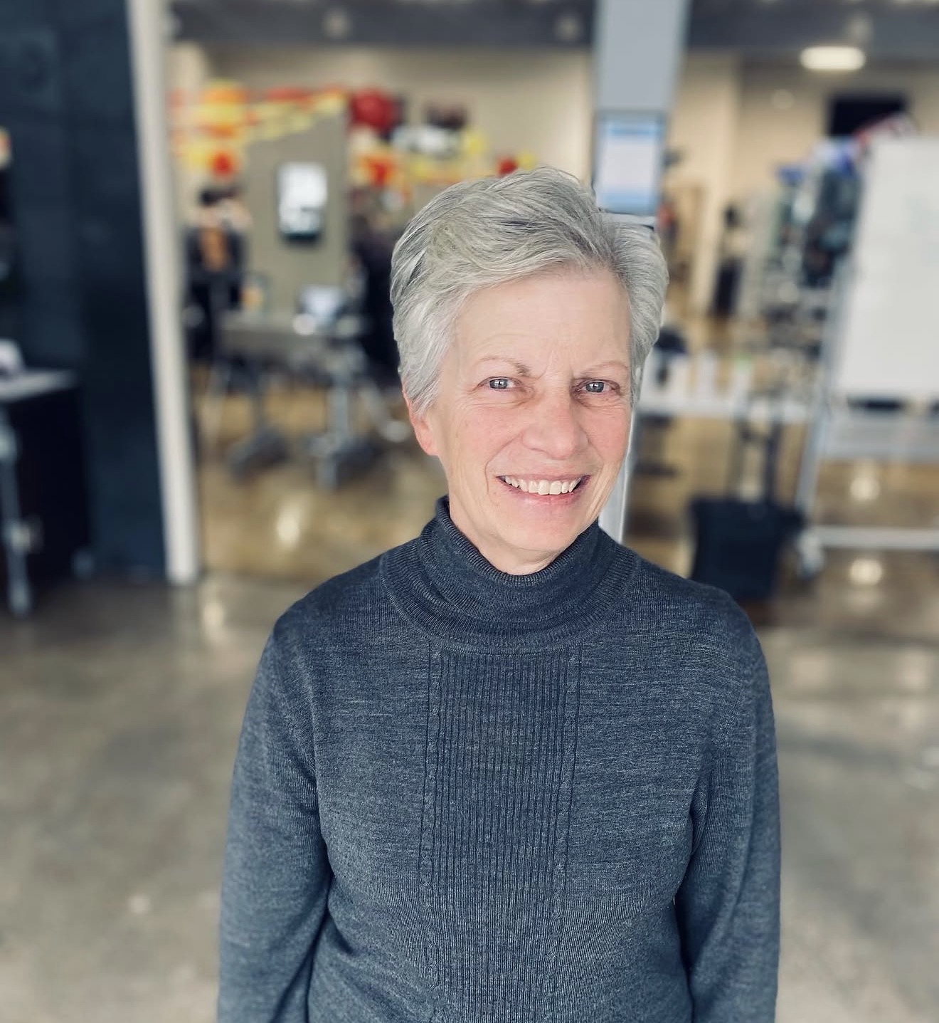 Smiling older woman with short gray hair wearing a dark gray turtleneck sweater, standing indoors in a store or warehouse.
