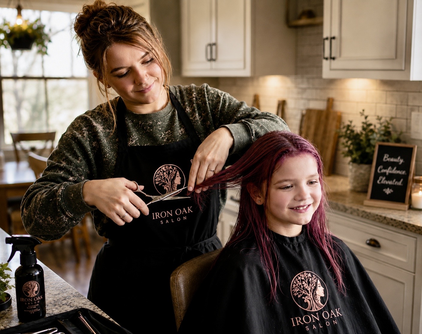 Woman cutting a young girl's hair in a kitchen, both wearing aprons, smiling, with kitchen cabinets and a window in background.