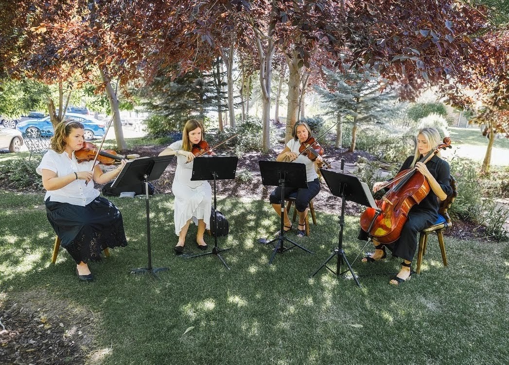 Four women playing string instruments outdoors under trees, seated on chairs with music stands, in a park setting.