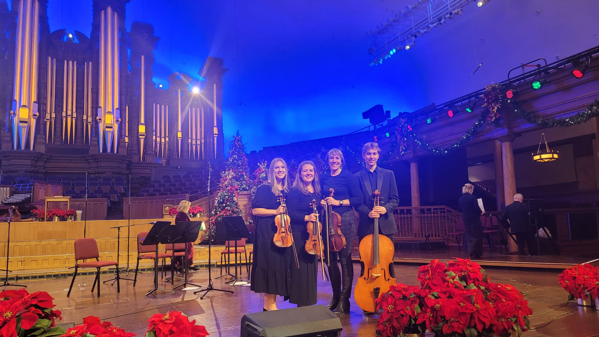 Four musicians with string instruments on stage in a concert hall, decorated with red flowers and Christmas trees.