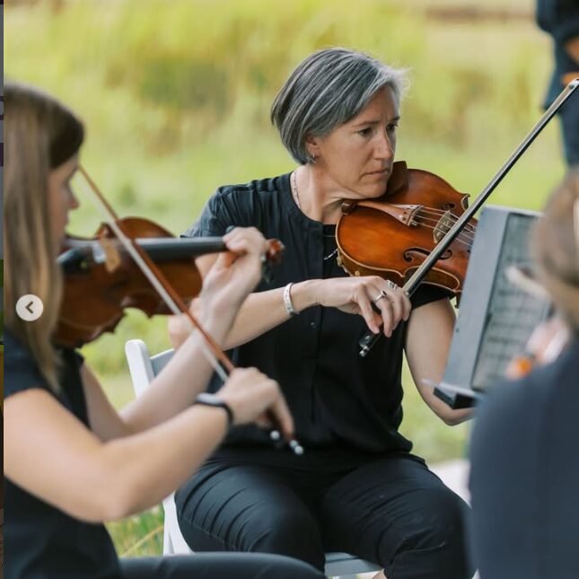 Woman with short gray hair playing violin outdoors with other musicians, blurred background of greenery.