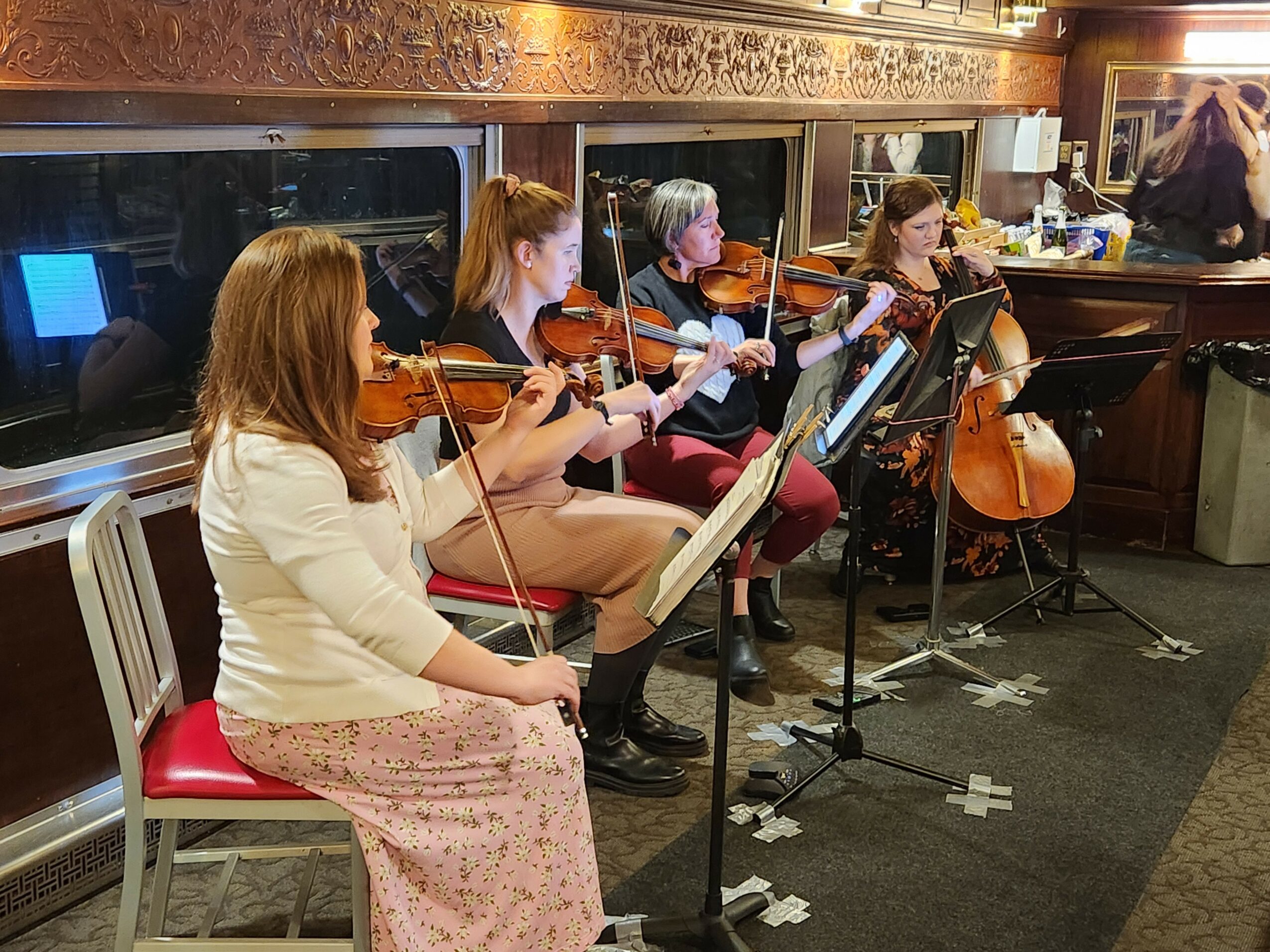 Four women playing violins and a cello in a room with a bar, music stands, and a window, seated in a row.