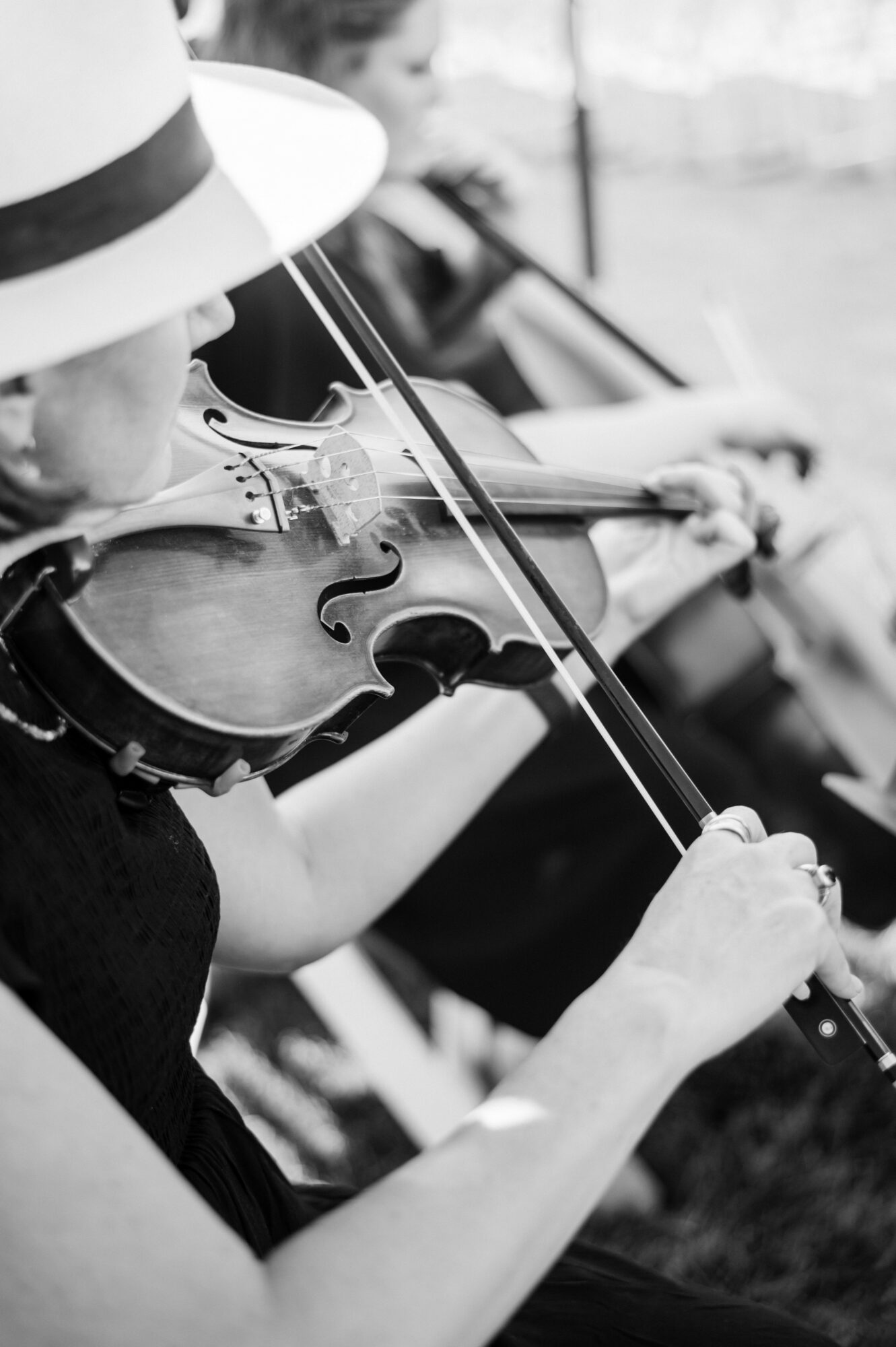 Person wearing a hat playing a violin outdoors, with another person in the background, in black and white.