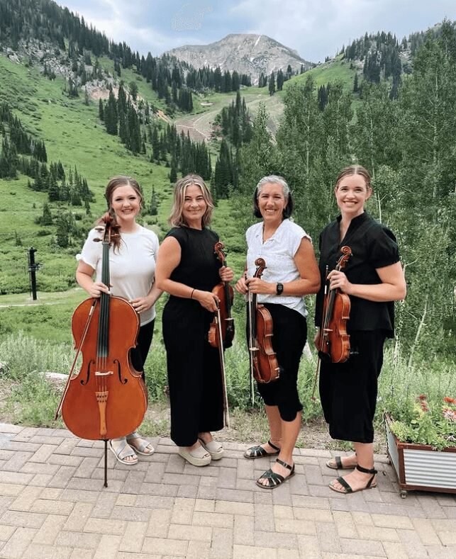 Four women holding string instruments outdoors in a mountainous landscape with green trees and a mountain in the background.