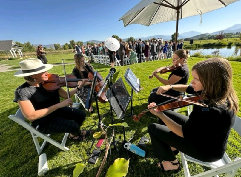 Four people playing violins outdoors near a pond, with an umbrella and a crowd in the background.
