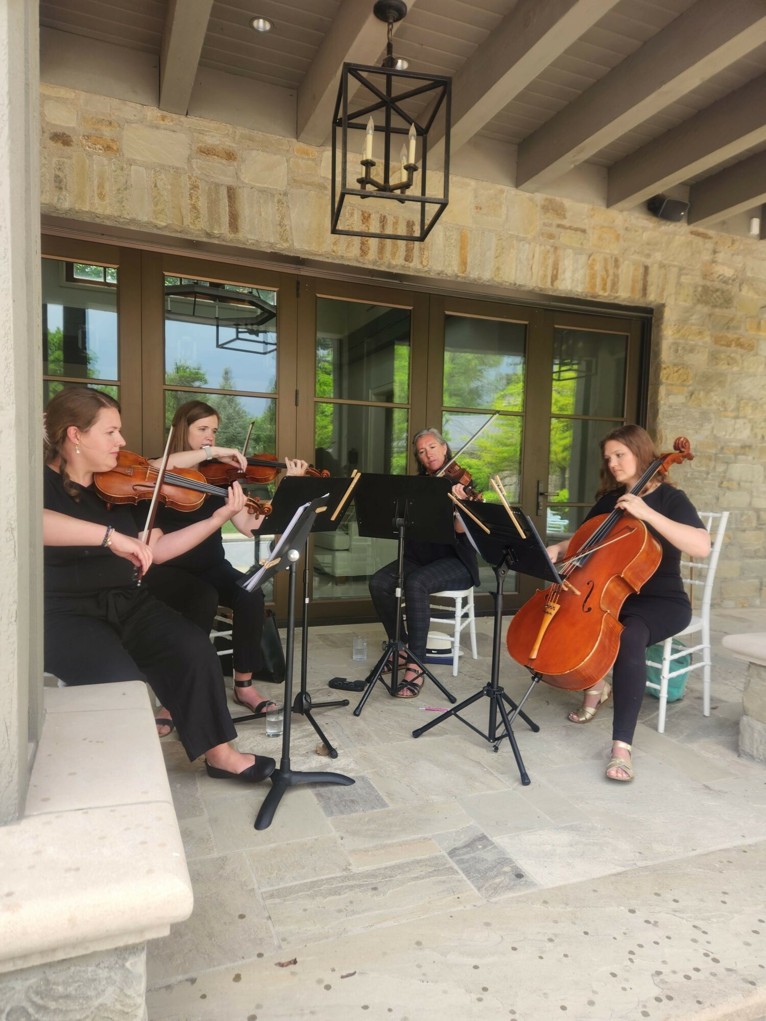 Three musicians playing string instruments outdoors near a building with large glass doors, with a woman standing nearby. Sunlight and greenery visible.