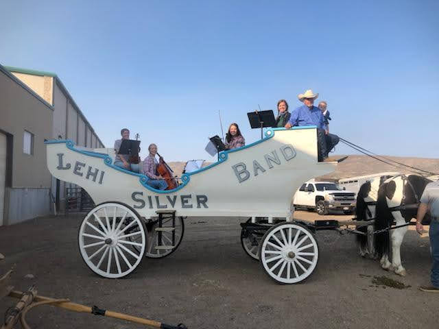 People sitting on a white horse-drawn carriage labeled 'Lehi Silver Band' with a clear blue sky background.