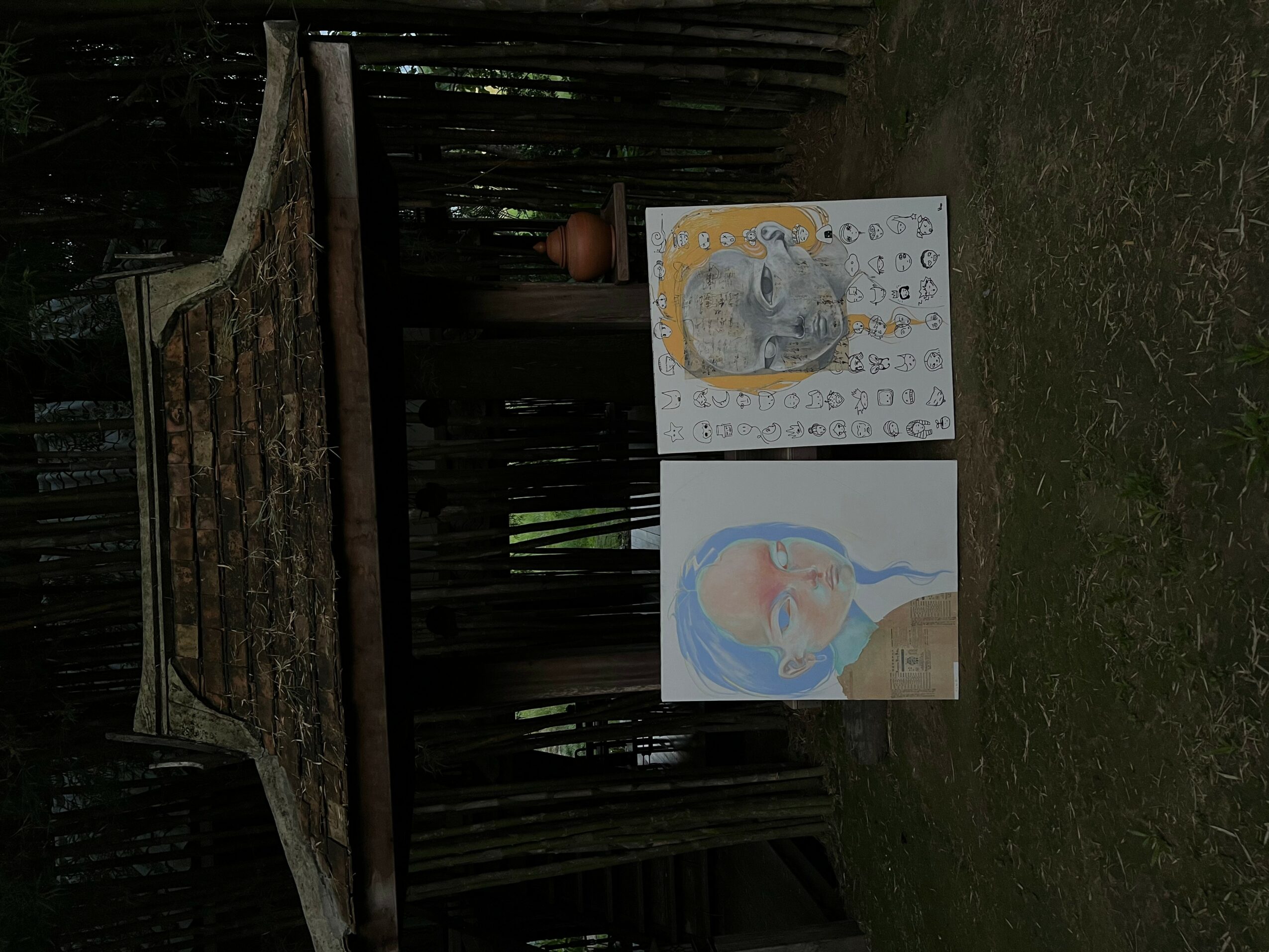 Two colorful drawings of animals on paper, placed on grass in front of a wooden fence, with a brick structure and potted plant in background.