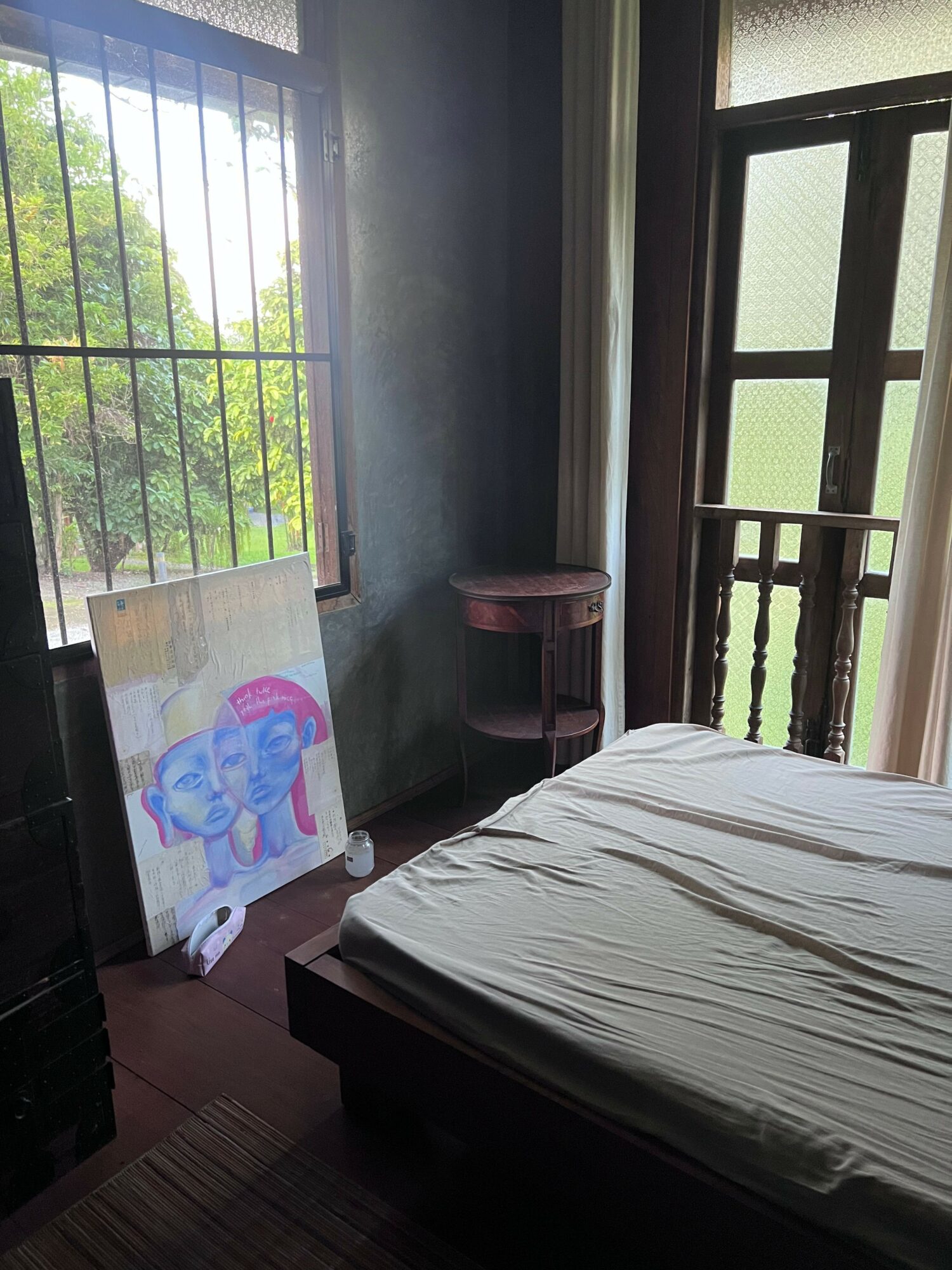 Bedroom with bed, window, and a colorful drawing on an easel, sunlight streaming through window bars.