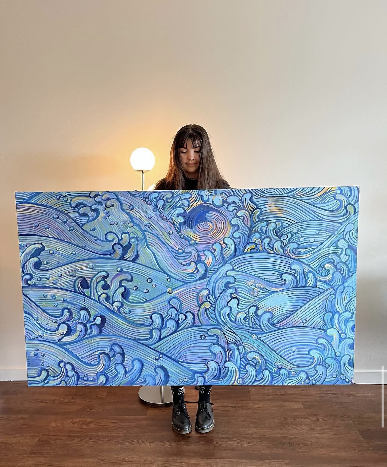 Person holding a large blue painting with swirling wave patterns, standing indoors with a lamp and plain wall behind.