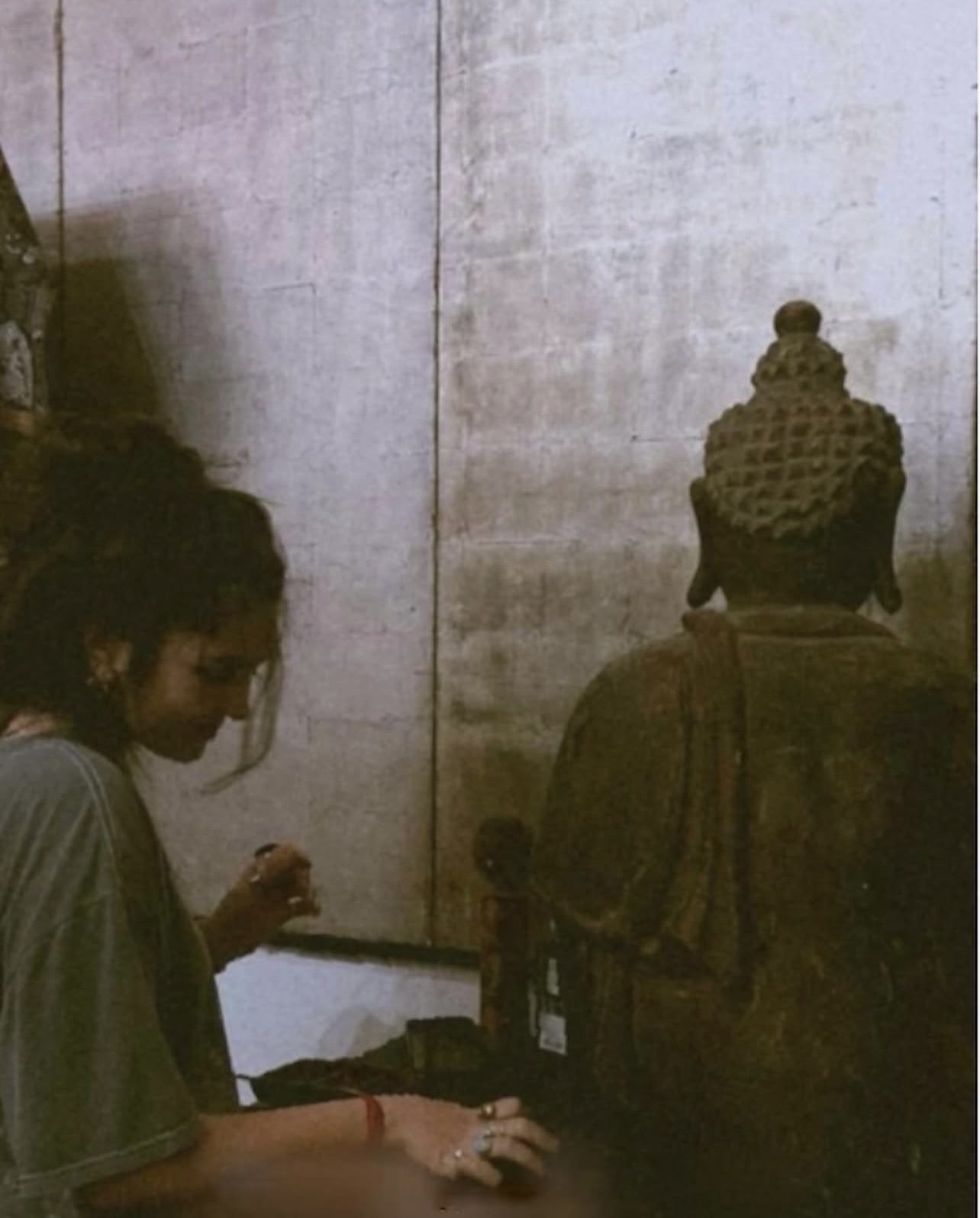 Two people sitting against a wall, one with a patterned head covering, facing away from the camera, in a dimly lit room.