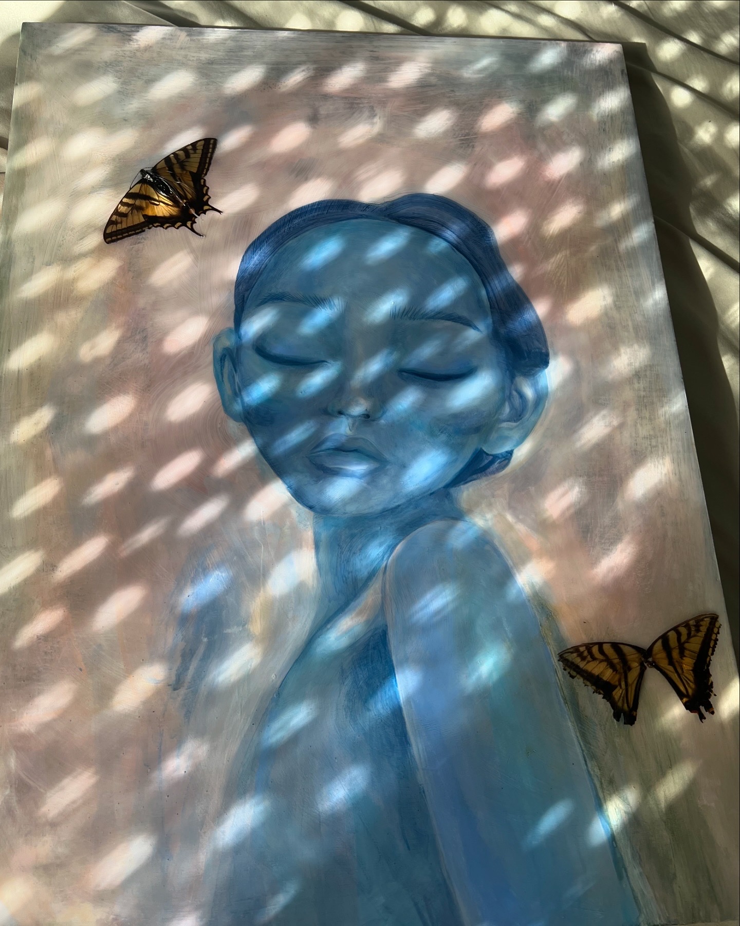 Framed artwork of a blue-toned face with closed eyes, surrounded by two butterflies, with light and shadow patterns on the surface.