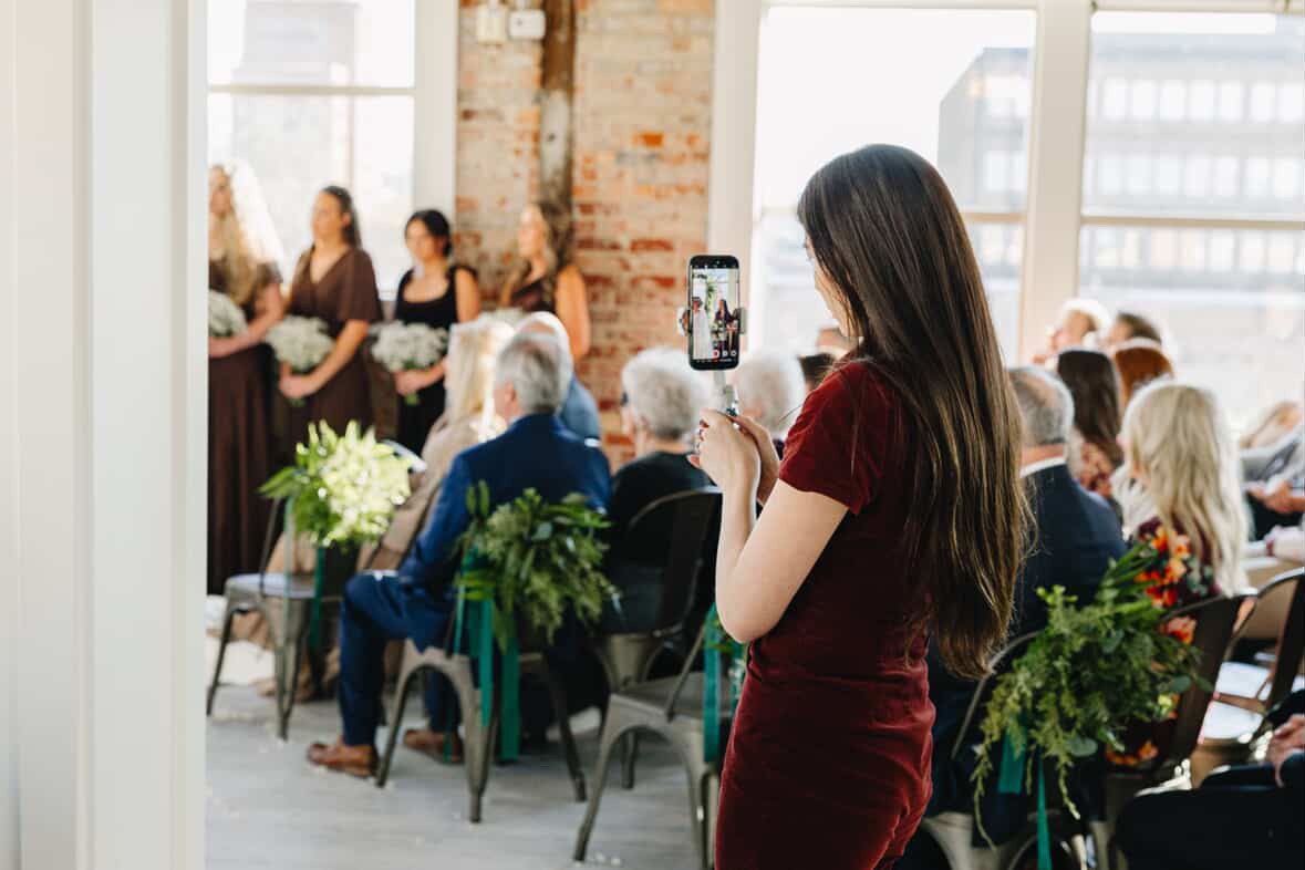 Woman in red dress taking a photo at an indoor event with seated audience and speakers on stage.