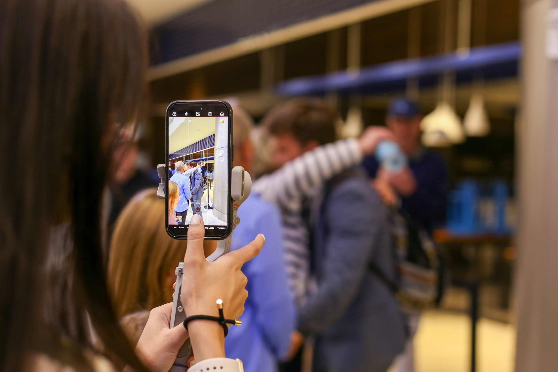 Person taking a photo with a smartphone of a group of people in a store or cafe, some wearing masks.