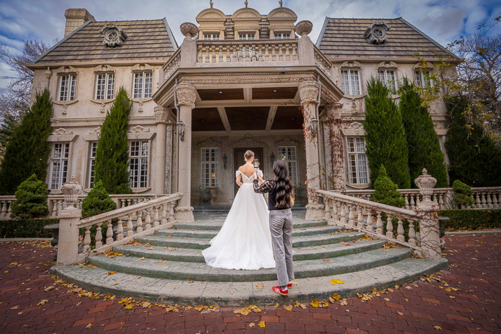 A bride and groom stand on steps in front of a large, ornate mansion with tall trees and a cloudy sky.